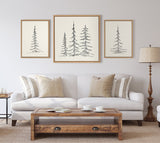 Three framed minimalist black ink evergreen tree sketches in varying sizes arranged above a beige sofa with neutral pillows in a light, cozy living room.