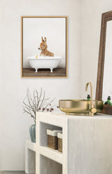 Framed canvas of a baby donkey in a white clawfoot bathtub, hanging on a beige wall above a modern bathroom counter with gold sink and faucet.