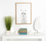 Framed black and white Sylvie Hairy Alpaca canvas print centered on white wall above a white table with green potted grass, stacked vintage books, white bird figurines, and white leaf sculpture.