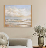 Framed painting of a beach scene with gentle waves under a cloudy sky in soft beige and blue tones hanging above a light gray couch and next to a table with a glass vase of dried flowers.