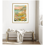 Framed landscape painting with coastal cliffs, wildflowers in the foreground, and a pastel sky, hung above a wooden bench with a draped cloth and open book.
