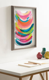 Framed abstract artwork featuring layered curved brushstrokes in pink, blue, green, and yellow hues with dotted accents, mounted on a white wall above a desk.