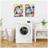 Two colorful abstract framed canvases with bold shapes and vibrant colors hang above a white front-load washing machine in a laundry room with a wicker basket and a white table with cleaning supplies.