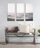 Three-panel canvas art showing calm ocean waves and sandy shore at sunset above wooden bench with textured white and gray pillows in neutral-toned room.