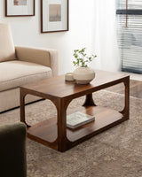 Rectangular wooden coffee table with rounded cutout corners and lower shelf, holding a textured ceramic vase with greenery and a small dish in a sunlit living room.
