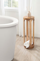 Tall, cylindrical Dahl wood drink table with vertical slats and a round top, holding a diffuser and rolled white towels, beside a white bathtub in natural light.
