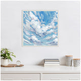Square framed canvas painting of a bright blue sky with swirling white and light purple clouds, hanging above a white desk with a small plant, books, and two patterned jars.