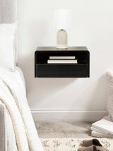 Black floating side table with rounded edges mounted on a white wall, featuring a beige lamp on top and books stored in an open shelf and drawer below.