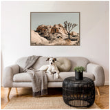 Framed canvas print of rugged tan rock formations and a Joshua tree under a gray sky, hanging above a light gray sofa with a round pillow and dog.