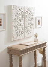Large two-panel white carved wood wall art with symmetrical floral and scroll patterns mounted above a light wood console table with carved apron and turned legs, flanked by two framed pictures on a white wall.