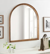 Wall-mounted arched wood frame mirror with a natural finish, reflecting window light in a bright room with framed black-and-white photos and a tabletop display.