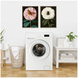 Laundry room with white front-loading washer centered between a wicker laundry basket and a white table holding folded towels, detergent, and a small plant, with two framed floral insect artworks above.