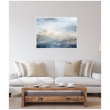 Abstract horizontal canvas with layered blue, beige, and gray tones evoking a serene oceanic scene, framed in white and hung above a light gray sofa with neutral pillows.