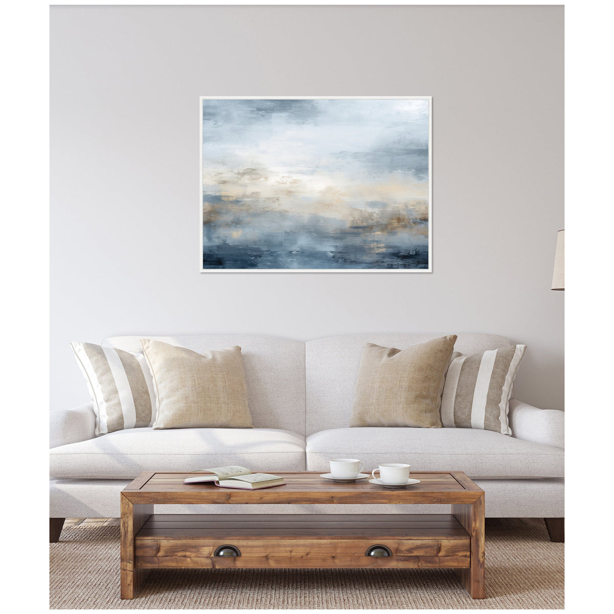 Abstract horizontal canvas with layered blue, beige, and gray tones evoking a serene oceanic scene, framed in white and hung above a light gray sofa with neutral pillows.