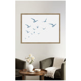 Minimalist wall art with blue silhouette seagulls flying across a white background in a wooden frame, above two dark green chairs and a round wooden table with a vase.