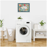 Framed wall art above a white front-loading washing machine shows a white cat surrounded by orange flowers and green leaves on a dark teal background.