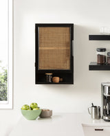 Black wood wall cabinet with woven rattan front door and open cubby shelf below, mounted on white wall above white kitchen countertop with green apples and coffee accessories.