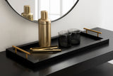 Matte black rectangular tray with gold handles holds gold cocktail shaker, matching golden tongs, and two dark smoky glass tumblers, placed on a black surface beneath a round mirror.