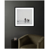 Framed print of two humpback whales surfacing with open mouths amid calm gray water, surrounded by small birds, hanging on a dark wall above an olive green armchair and black side table.