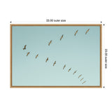 Framed canvas of a diagonal row of pelicans flying against a clear pale blue sky, measuring 33 by 23 inches in a light wood frame.