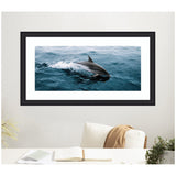 Framed artwork showing a dolphin leaping and creating splash detail in dark and light blue ocean waves, displayed above a white couch with a tan blanket.