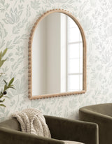 Arched wall mirror with a light wooden frame featuring rounded bead detailing along the edge, hung on a pale floral wallpapered wall above olive-green velvet chairs.