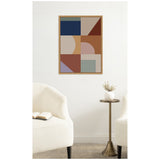 Framed abstract art with six rectangular sections featuring geometric shapes in muted blue, beige, brown, and lavender, above a simple round brass side table with a white vase and dried flowers.