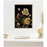 Framed canvas artwork of pale and dark peach flowers with green stems on a black background, hung above a white desk with neutral decor and dried grasses.