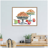Watercolor painting of two apple pies on blue pedestal dishes surrounded by red and green apples with stems and leaves, framed and hung on a white wall above a wooden desk.