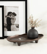 Oval dark brown decorative tray with four short legs holding a small matte black vase with delicate white flowers, set against a white surface with framed black-and-white photo.