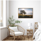 Coastal landscape painting featuring a lone tree on a grassy shore under a partly cloudy sky, framed in wood and hung on a white wall above natural wicker chair.
