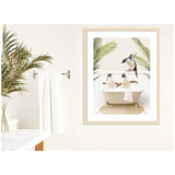 Wall art print featuring two penguins sitting in a vintage clawfoot bathtub with palm fronds overhead, displayed in a light wood frame on a white textured wall.