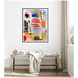 Framed abstract canvas with bold colorful shapes including black, blue, red, coral, and olive green, hanging above a natural wood bench with a white cloth and open book in a minimalist room.