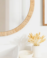 Partial view of a round wall mirror with a textured light wood frame above a white surface holding a beige vase with wheat-like dried grasses and a shallow dish.