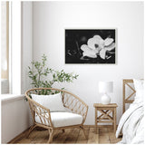 Black and white close-up photograph of a magnolia flower with soft petals and a dark background, framed and hanging above a cozy wicker chair with white cushions beside a wooden nightstand and lamp.