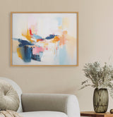 Abstract coastal-inspired artwork with soft pastel blues, pinks, yellows, and navy brushstrokes in a light wood frame, hanging above a beige sofa and floral vase.