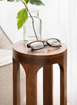 Round wooden side table with smooth edges and a rich brown finish, topped with a clear glass vase holding a green leafy branch and a pair of black-framed eyeglasses.
