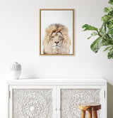 Framed portrait of a lion's face with a full mane on a white background, hanging above an ornate white carved cabinet beside a white vase and green plant.
