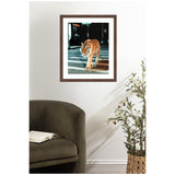 Framed photograph of a tiger walking across a city street at night, featuring black background and bright tiger stripes, hung on a white wall above a green armchair and potted plant.