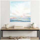 Abstract seascape painting with soft pastel blues, pinks, and whites suggesting water and sky, framed in white and hung above a wooden table with books, a mug, and a small vase.