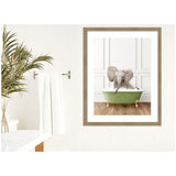 Framed wall art showing a baby elephant sitting in a green clawfoot bathtub against a white paneled wall, displayed in a minimalist bathroom setting with white towels and a plant.