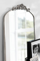 Tall arched mirror with ornate carved floral crest and a distressed white frame, reflecting a blurred window and framed black-and-white photograph beside it.