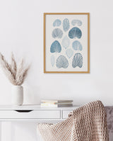 Framed art print of abstract blue seashell shapes arranged in a grid on white wall above white desk with books and a vase of dried pampas grass.