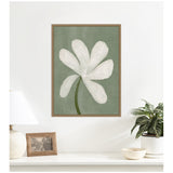 Framed canvas print of a white flower with six rounded petals and a green stem on a muted green background, hanging above a white surface with a lamp, photo frame, plant, and bowl.