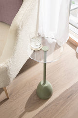 Small round drink table with clear acrylic top and matte olive-green metal base, positioned next to a textured cream armchair on light wood flooring near a curtained window.