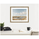 Framed photograph of a marshland with golden grasses, water pools, and a blue sky with scattered clouds, mounted above a white desk with books, a black mug, and a plant.