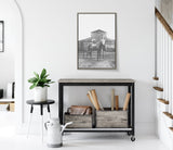 Black and white framed canvas of a horse standing in front of a barn, hung above a rustic console table with wooden crates and rolled papers, accompanied by a potted plant and watering can.