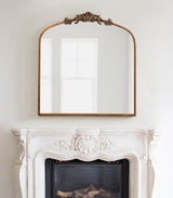 Rectangular arch mirror with a carved wooden frame and ornate leaf design at the top, hanging above a detailed white fireplace mantel.