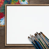 Blank 30x30 framed canvas resting on a wooden surface with a colorful painter's palette and a cluster of paintbrushes arranged at the lower right corner.