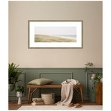 Framed coastal landscape print above a wooden bench with a green cushion and beige throw, surrounded by potted plants against beige and sage green paneled walls.